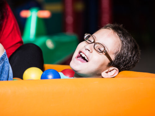 Close de um menino de aproximadamente 8 anos, cabelos curtos, óculos e largo sorriso. Está na piscina de bolinhas e à sua frente 3 bolas nas cores amarelo, azul e vermelho