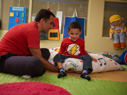 Um menino de aproximadamente 7 anos, calça jeans, meias e camiseta vermelha, sorri, sentado numa grande almofada branca com estampa colorida. Sentado ao seu lado, um jovem senhor, calça preta e camiseta vermelha, com a mão direita embaixo da almofada. Ao fundo, brinquedos