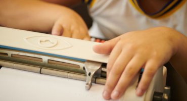 Detalhe de uma máquina braile, com as mãos de um menino, sendo que a mão direita toca uma das teclas, enquanto a esquerda, faz a leitura no papel