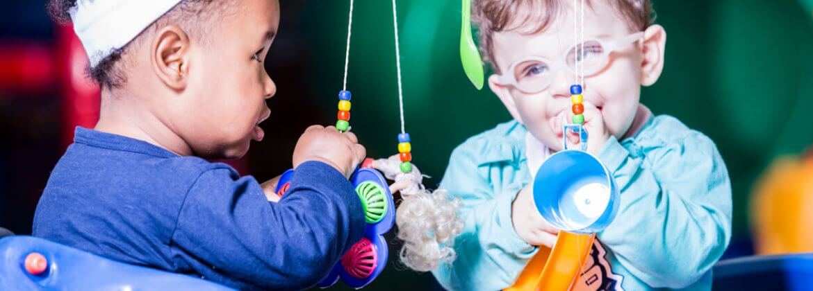 Dois bebês, brincando com um móbile, cujo os objetos pendurados nele são, uma colher verde, um chocalho roxo, uma boneca pequena e uma caneca azul.