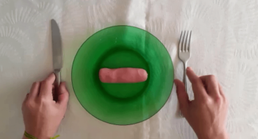 Prato na cor verde no centro da imagem. No prato há uma massinha de modelar representando uma salsicha. À esquerda mão segurando uma faca e à direita mão segurando um garfo
