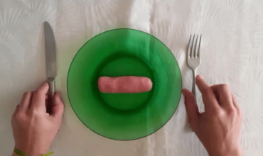 Prato na cor verde no centro da imagem. No prato há uma massinha de modelar representando uma salsicha. À esquerda mão segurando uma faca e à direita mão segurando um garfo