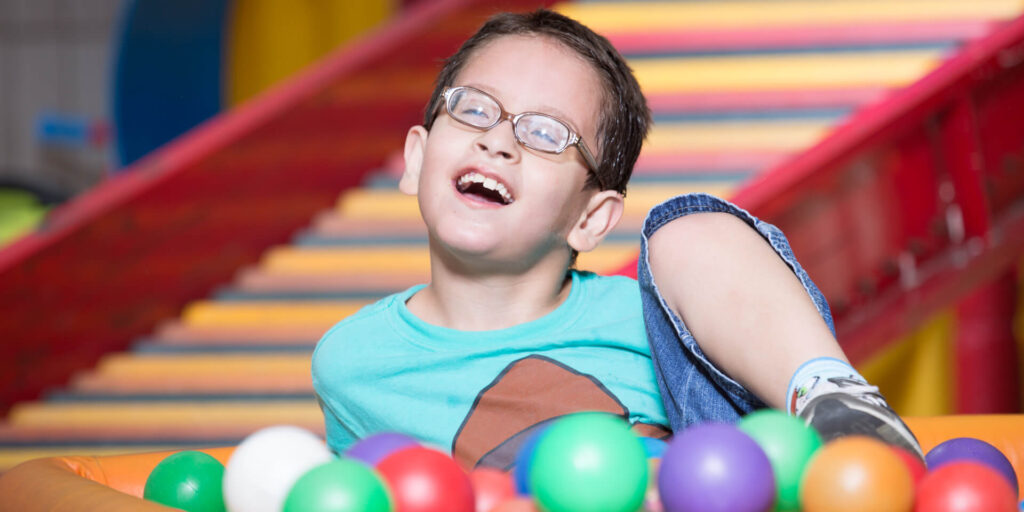 Close de um menino de aproximadamente 8 anos, cabelos curtos, óculos e largo sorriso. Está na piscina de bolinhas e à sua frente bolas em cores variadas