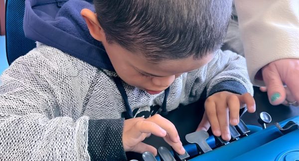 Criança concentrada digitando em uma máquina de escrever Braille azul. Ela usa um casaco cinza com capuz e posiciona os dedos sobre as teclas do equipamento. Ao fundo, uma pessoa adulta com casaco claro apoia a atividade.